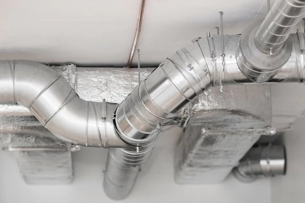 Close-up of silver metal ventilation ducts and pipes with insulation material in a modern building's ceiling. The ducts are interconnected with brackets and clamps, forming a network for air circulation. The ceiling is white with a smooth finish.