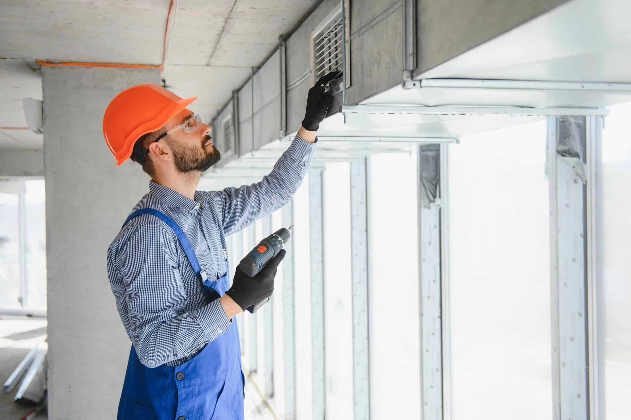 A construction worker in a blue overall and orange helmet uses a power drill to install or repair an air vent. He is working on ductwork in a building under construction. Natural light streams through large windows.