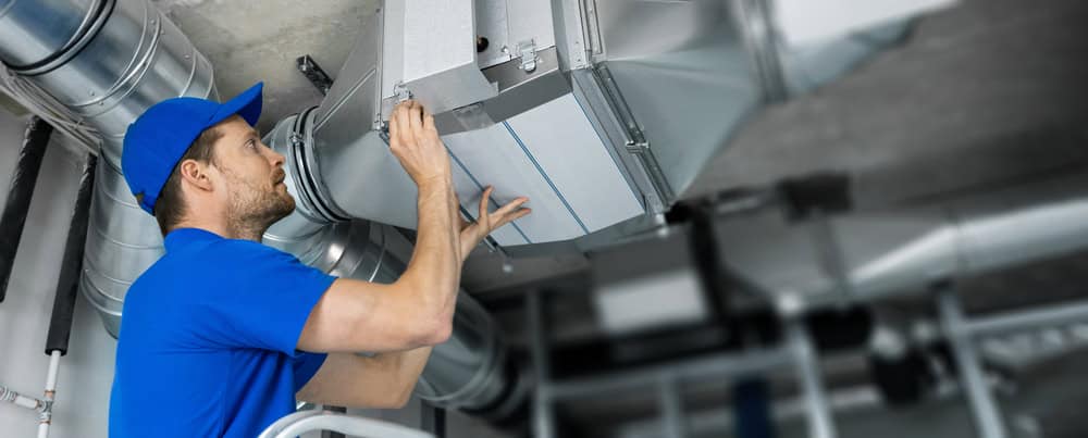 Technician in a blue uniform and cap works on a large metal ventilation duct in an industrial setting, adjusting or repairing the system from a ladder. Industrial piping and ducts are visible in the background.
