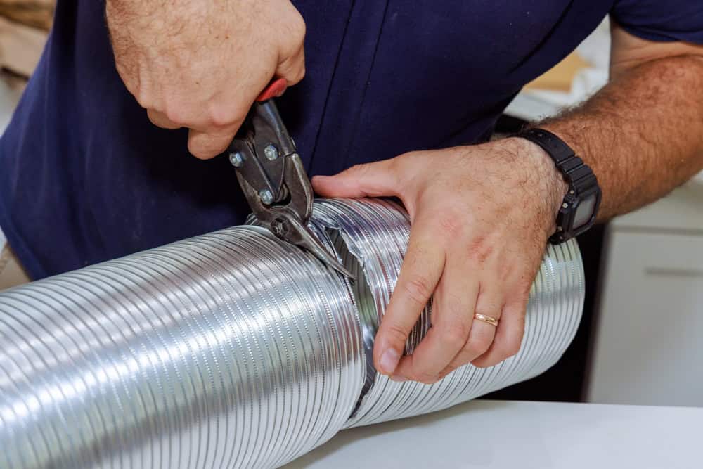 A person wearing a dark shirt uses tin snips to cut a flexible metallic duct. The person wears a black watch and a gold ring on their left hand. The duct is positioned on a white surface.