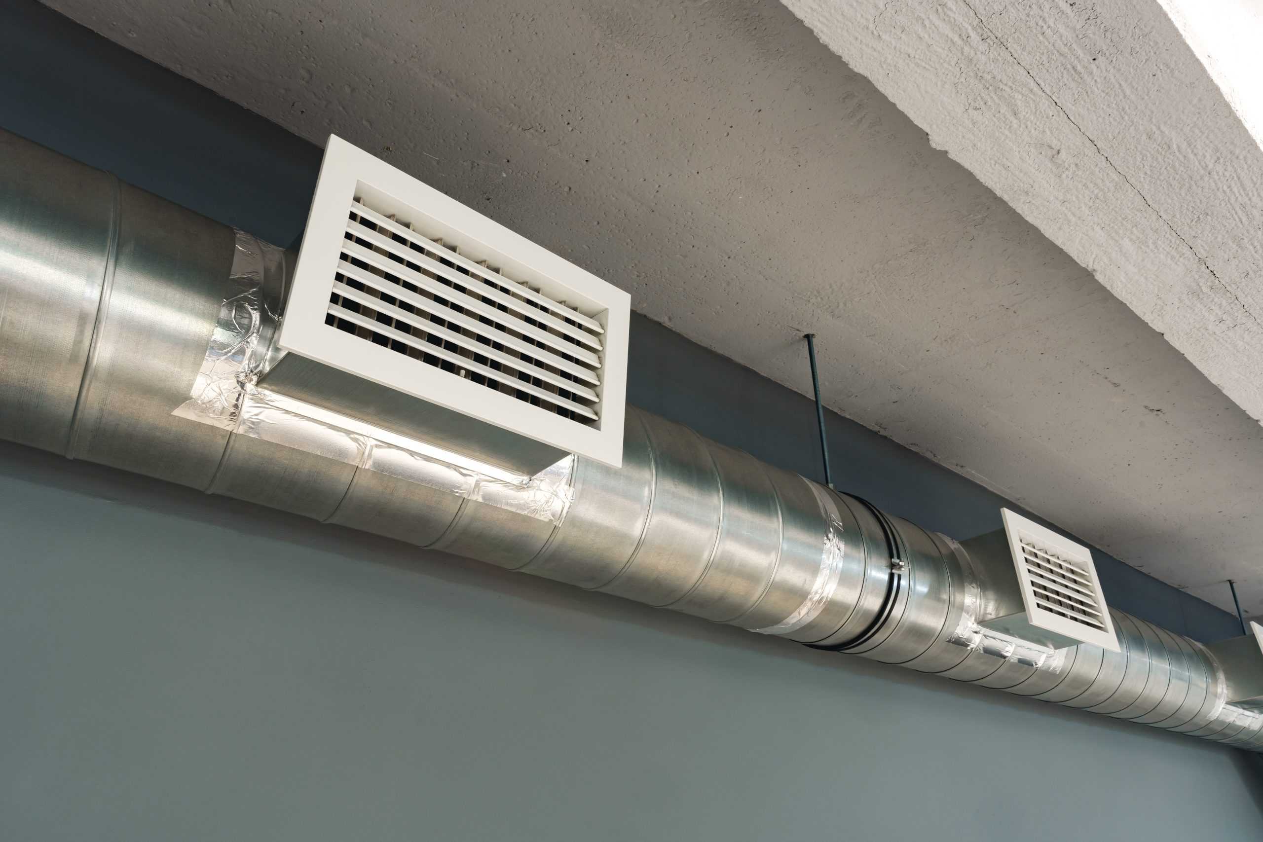 A metal air duct system with rectangular vents is mounted on a ceiling. The duct runs horizontally, and the background shows a gray wall. The ceiling has a rough, textured surface.