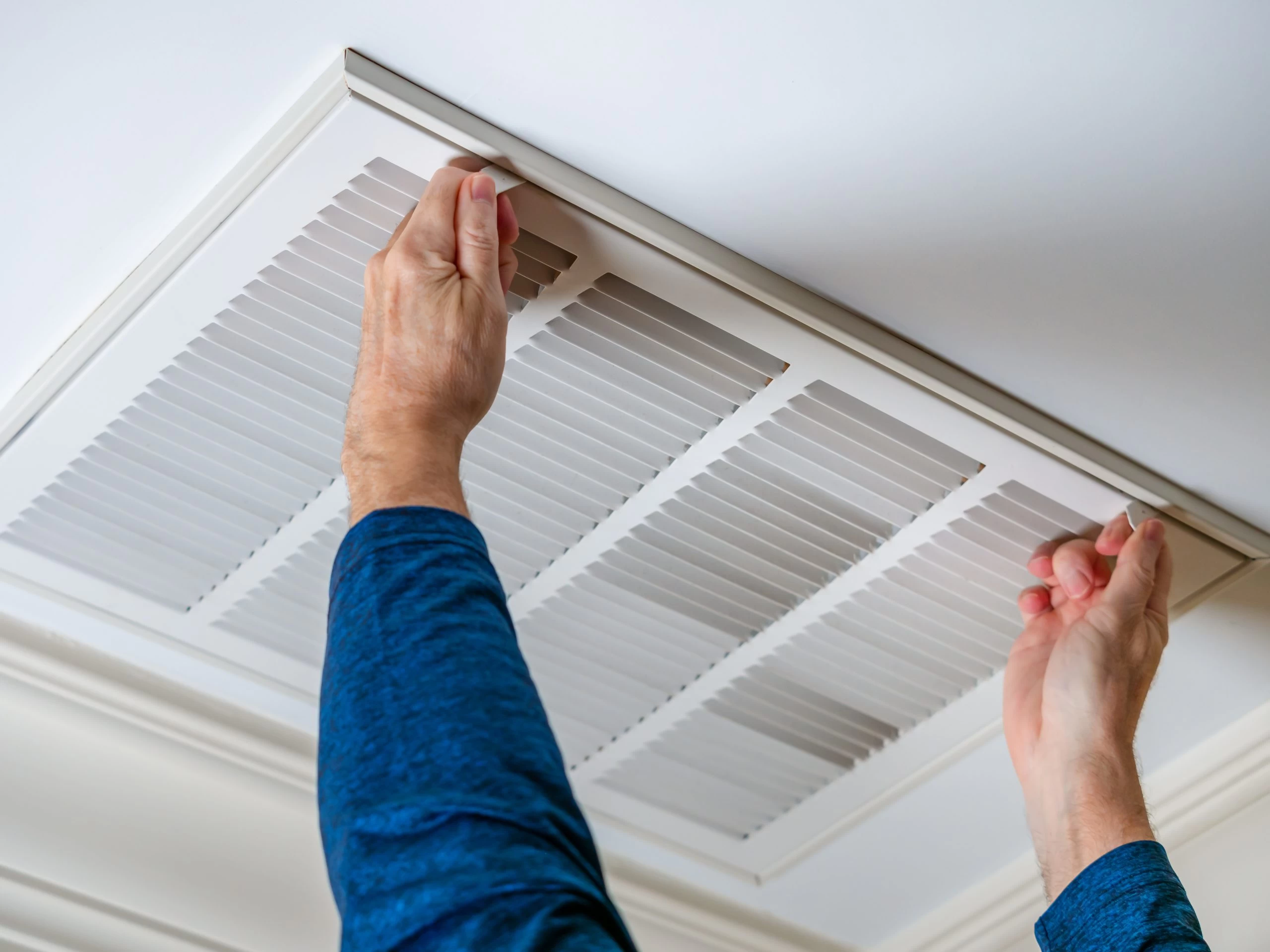 Person in a blue long-sleeve shirt adjusting or installing a white ceiling air vent. The hands are firmly gripping the vent cover, which has horizontal slats. The background shows a white ceiling.
