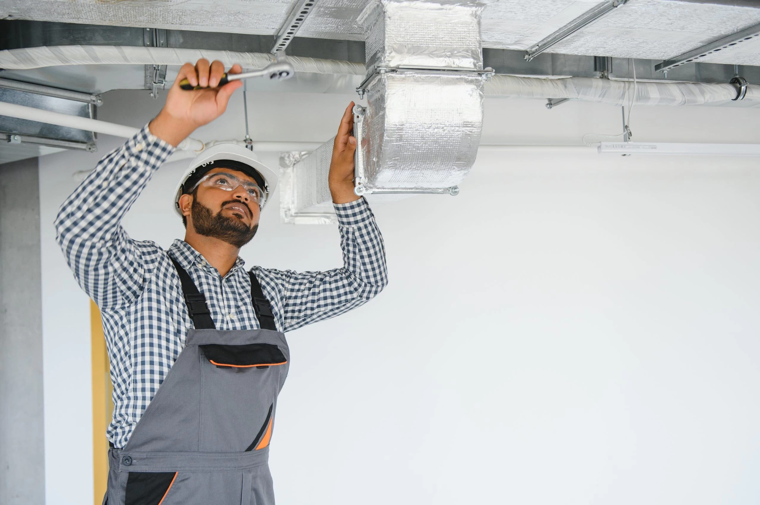A person wearing a hard hat and safety goggles is using a screwdriver to work on a metallic duct system. They are dressed in a checkered shirt and gray coveralls, standing in a room with a white wall and exposed ceiling pipes.
