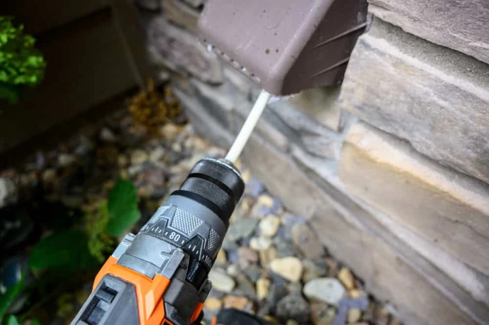 A drill with an orange and black handle is inserting a white cable through a small hole in an outdoor wall. The wall is made of brick-like stones. Green plants and small rocks are at the base of the wall.