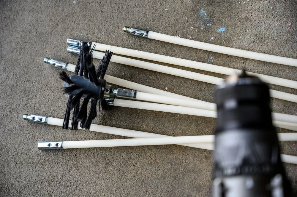Close-up of chimney cleaning rods with metal connectors and a black brush on concrete. A blurred object resembling a drill is in the foreground, suggesting they are tools for cleaning or repair.