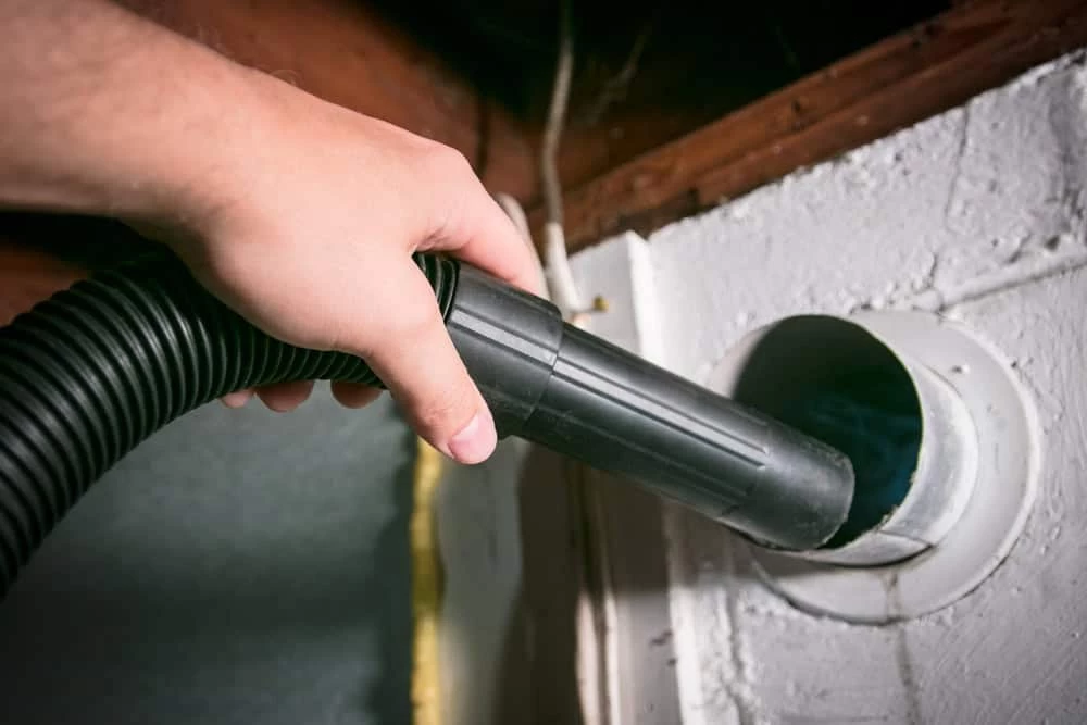 A hand holding a vacuum hose is inserted into a round duct on a white wall. The hose is black, and the surrounding area includes a wooden beam and part of an insulated wall.