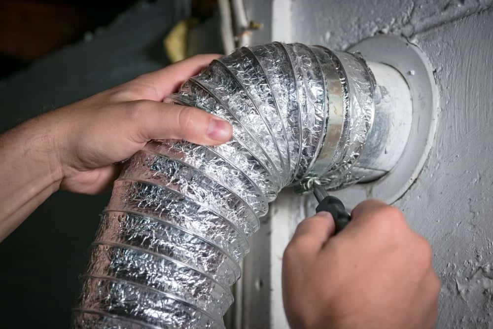 A person uses a screwdriver to secure a flexible, metallic dryer vent hose to a wall connector. The background is a plain, painted wall. The person's hands are visible, focusing on tightening the clamp around the hose.