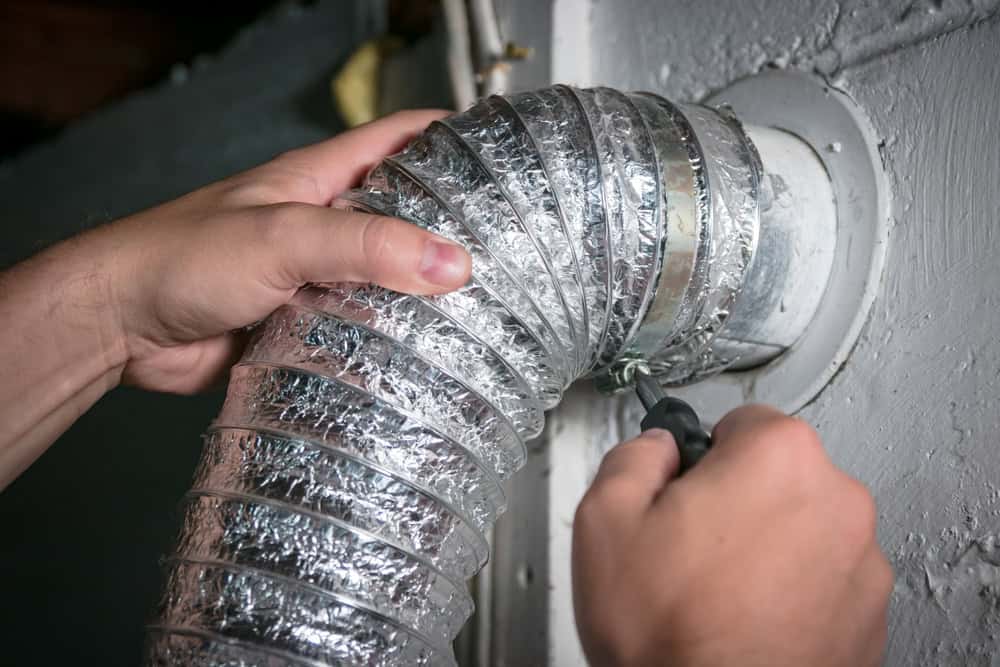 A person uses a screwdriver to secure a flexible, metallic dryer vent hose to a wall connector. The background is a plain, painted wall. The person's hands are visible, focusing on tightening the clamp around the hose.