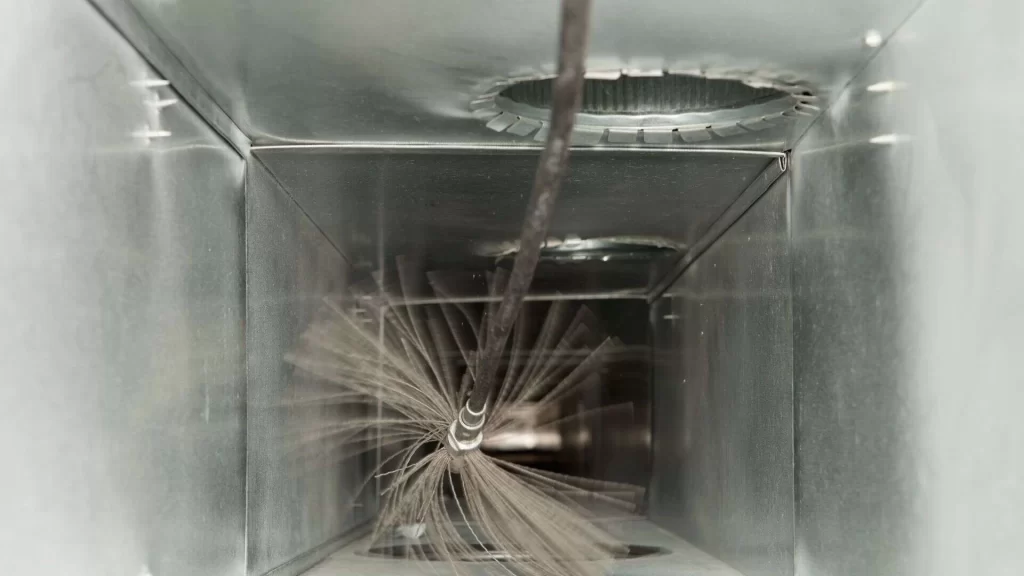 A round brush attached to a long rod is cleaning the inside of a metal air duct, with dust particles visible around the brush bristles.