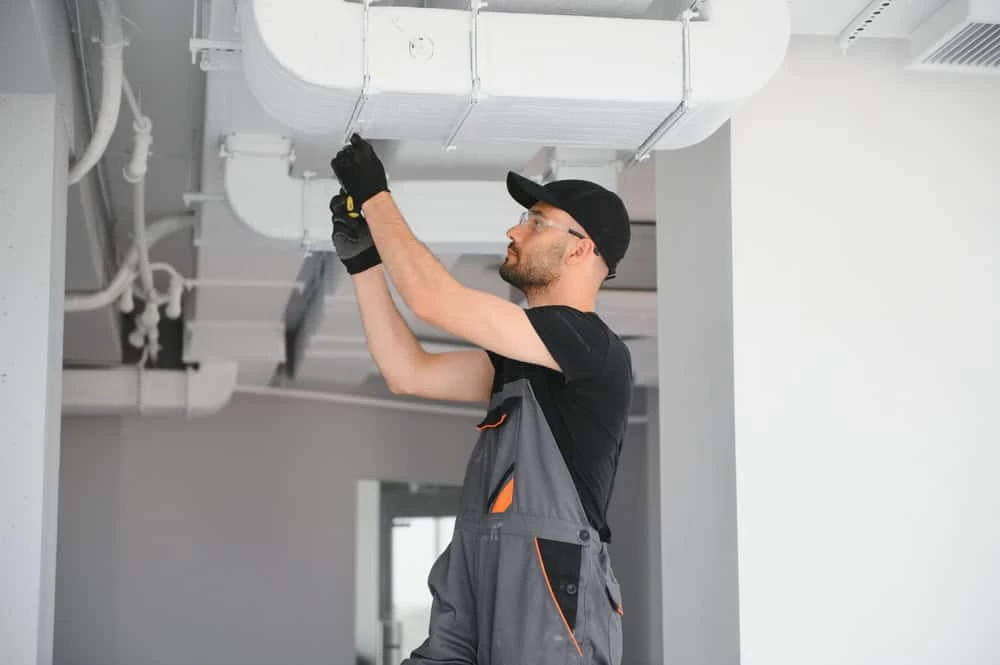A worker wearing gloves, safety glasses, and a cap installs or repairs ductwork on a ceiling in a modern, industrial-style building.