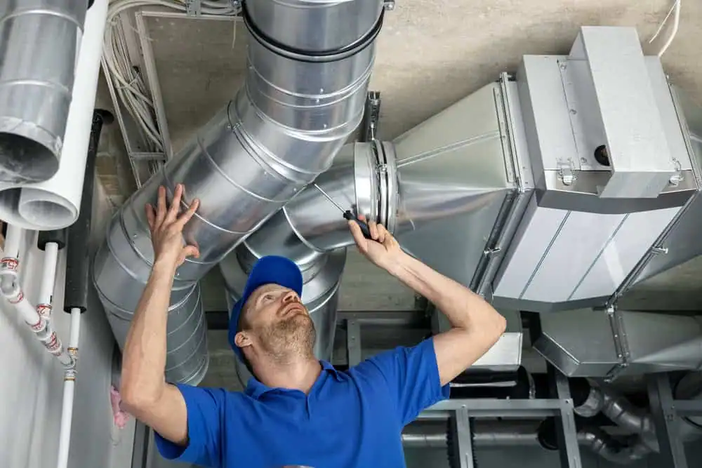 A technician in a blue shirt and cap uses a screwdriver to adjust metal air ducts on a ceiling in an industrial or commercial setting, ensuring quality Air Duct Cleaning Sacramento residents can trust.