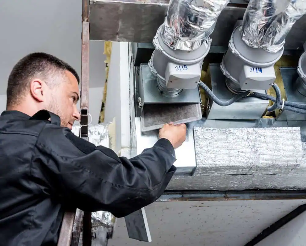 A technician in a black uniform inspects an air duct system, adjusting a vent or filter beneath large, insulated pipes and metal components on a ceiling—showcasing expert air duct cleaning in Contra Costa County, CA.